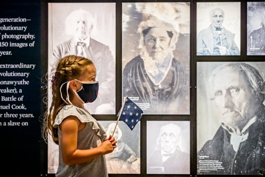 A young, female visitor, wearing a mask and holding a 13-star flag, admires photographs in the Museum's galleries.
