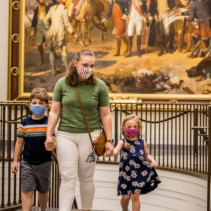 A masked family walks in front of The Siege of Yorktown painting located outside the galleries.