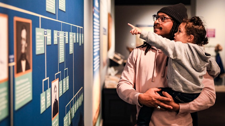 A father holds his child as they look at the Forten family tree in the Museum's Black Founders exhibit.