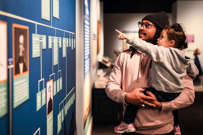A father holds his child as they look at the Forten family tree in the Museum's Black Founders exhibit.
