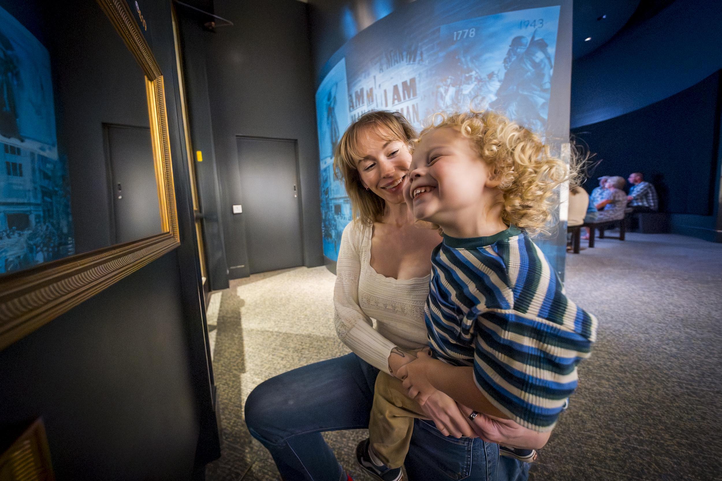 A woman and child smile into a mirror with an exhibit behind them