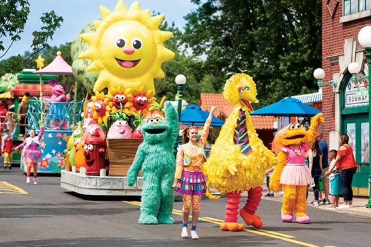 This image shows Big Bird leading a parade of Sesame Street characters and floats at Sesame Place.