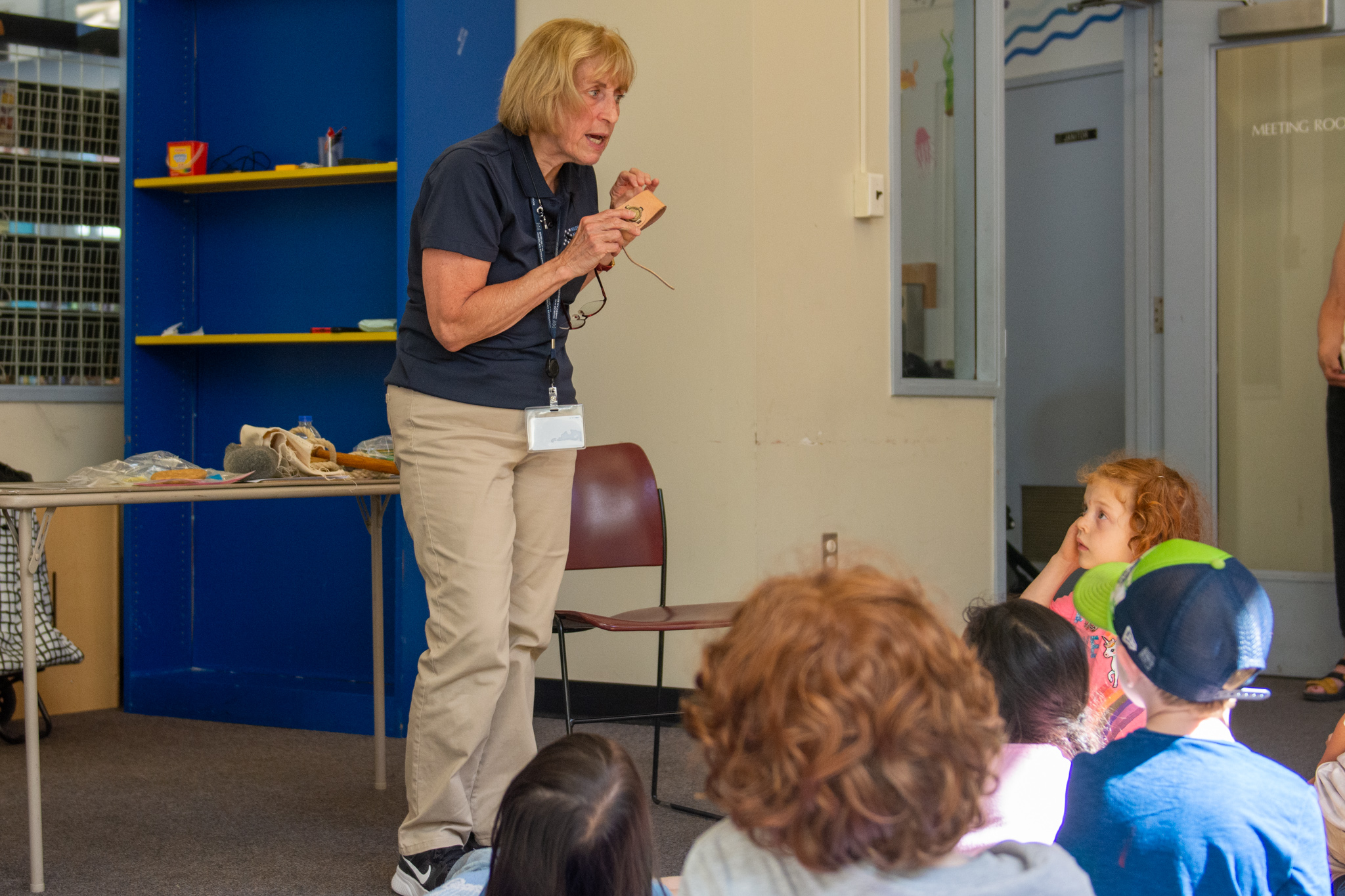 Woman with light blonde hair, navy polo, and khakis speaks to group of children