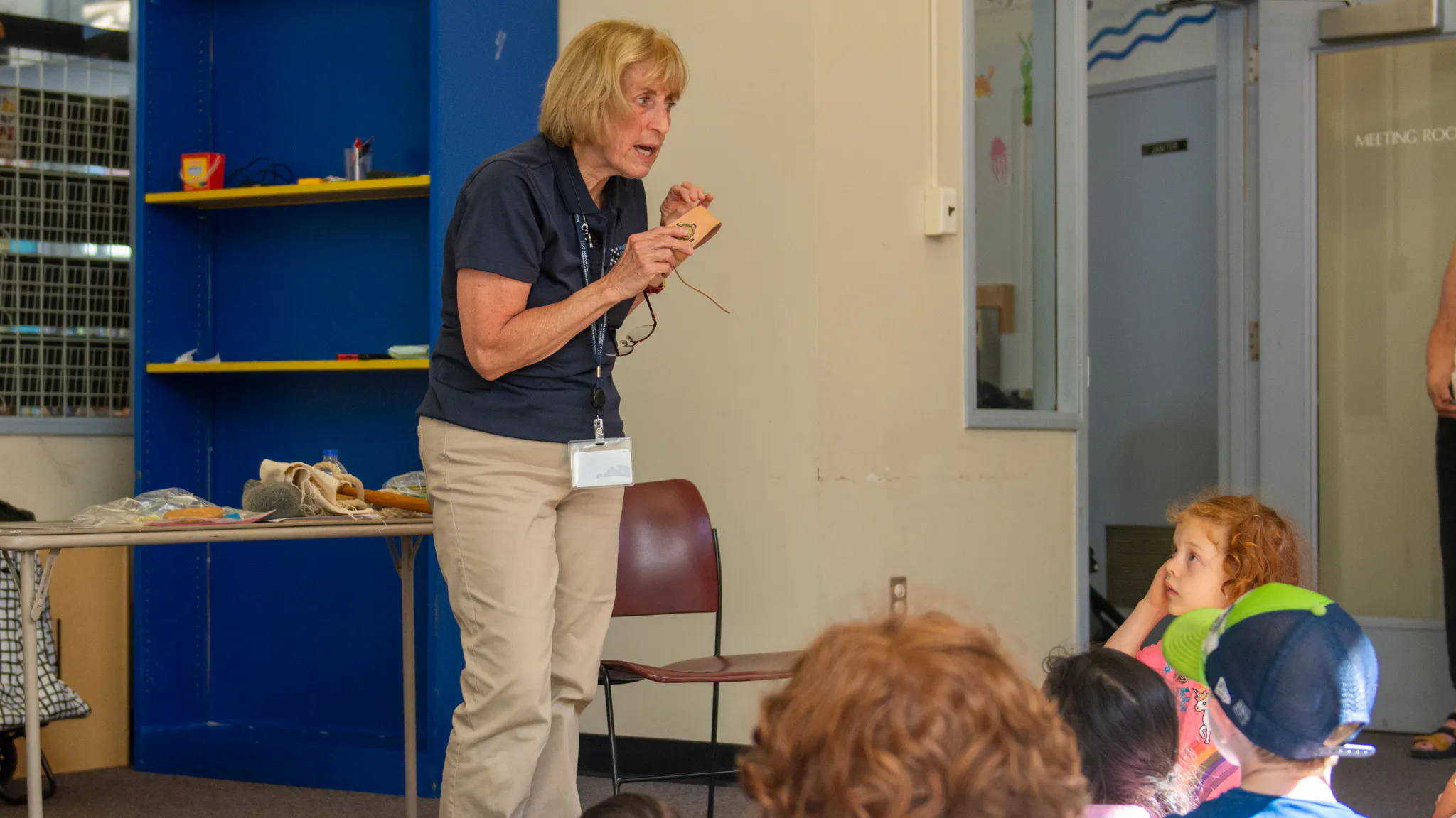 Woman with light blonde hair, navy polo, and khakis speaks to group of children