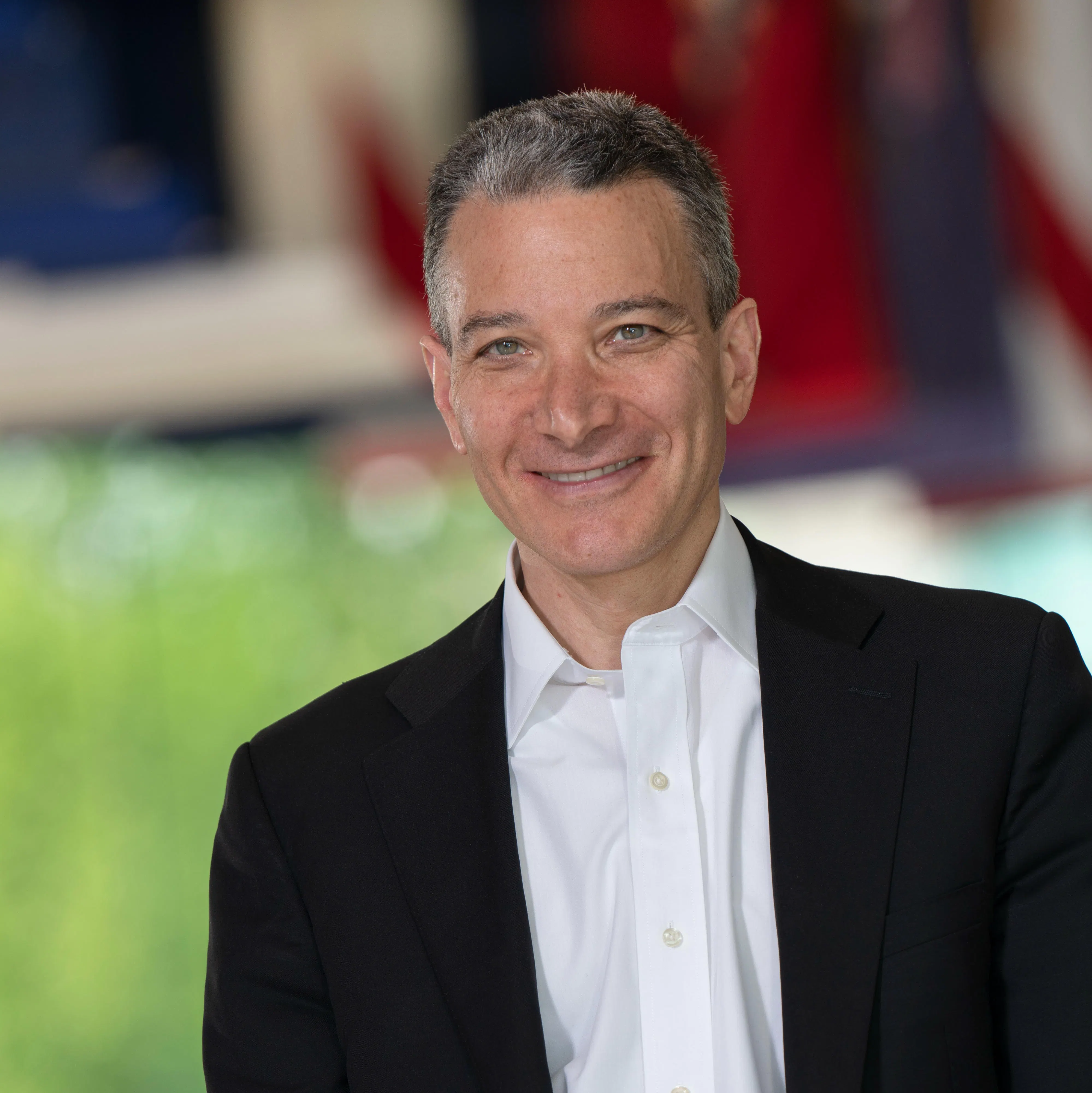 A man with grey hair, a white dress shirt, and black jacket smiliing at the camera