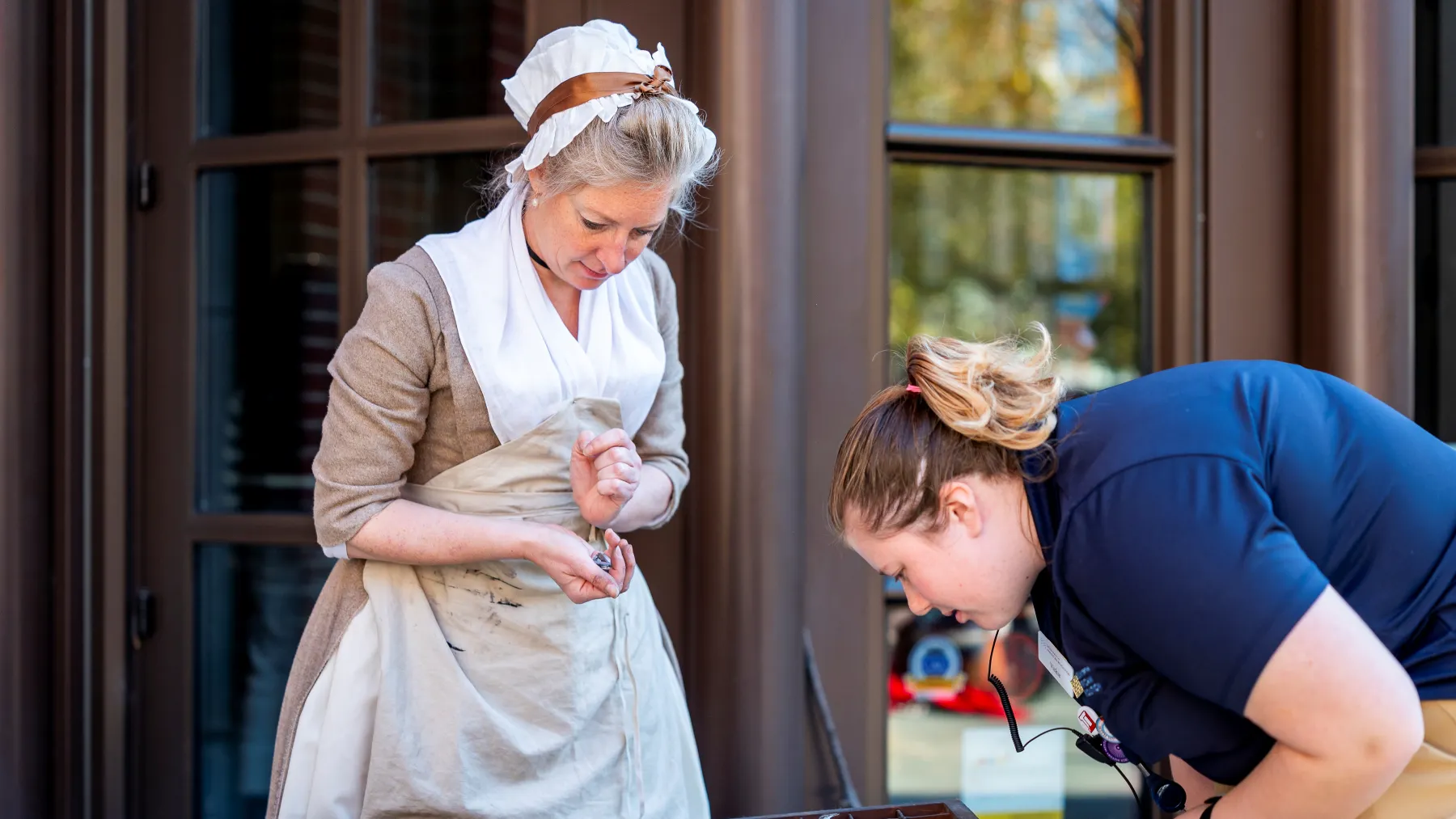A woman in 18th century dress stands behind a table of typesetting implements while a staff member looks at them