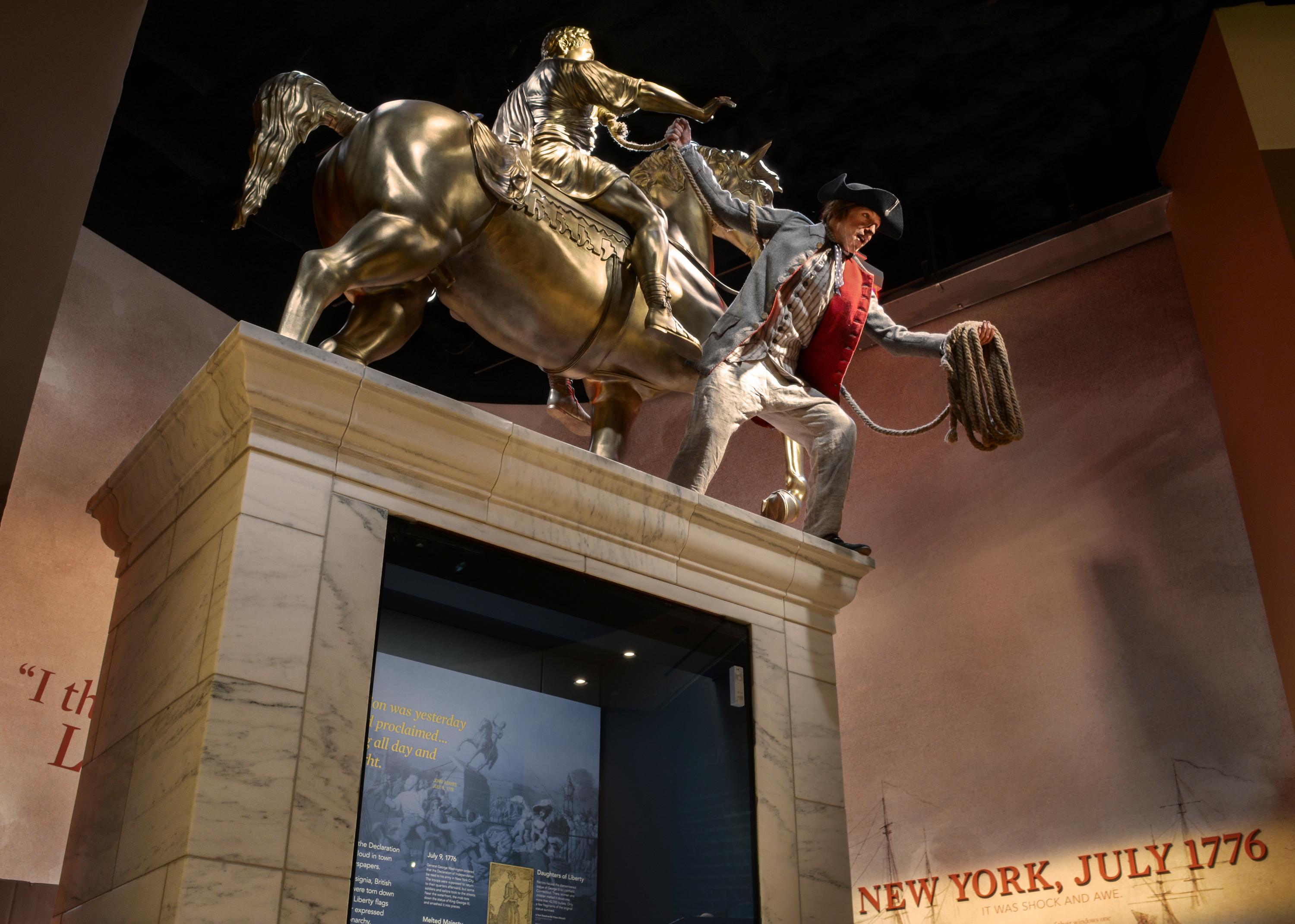 A life size figure dressed in 18th century clothing pulls down a statue of King George III