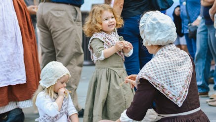 A young girl dressed in 18th century clothing talks to an adult living history interpreter on the Museum's plaza during Occupied Philadelphia.