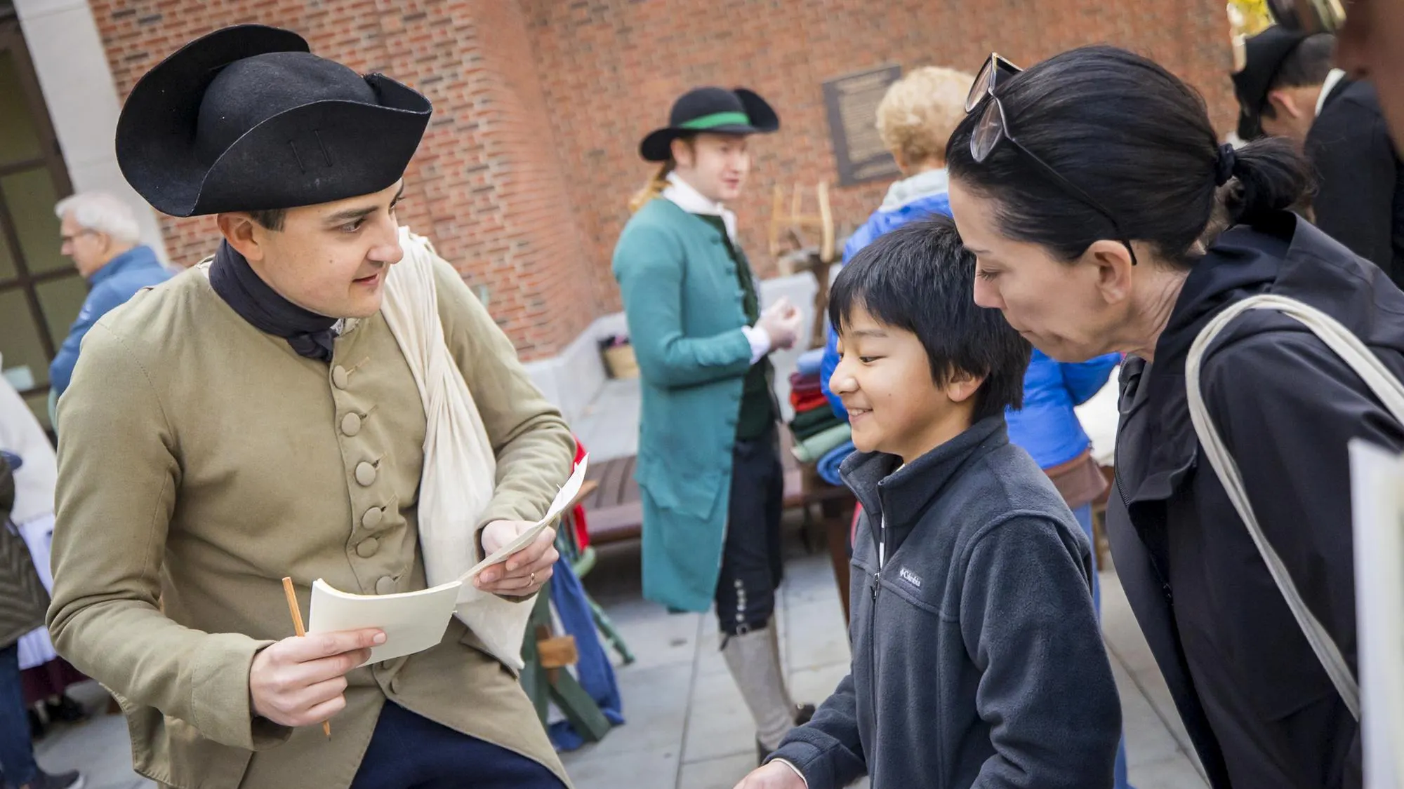 A costumed interpreter speaks with a woman and child while both hold pieces of paper
