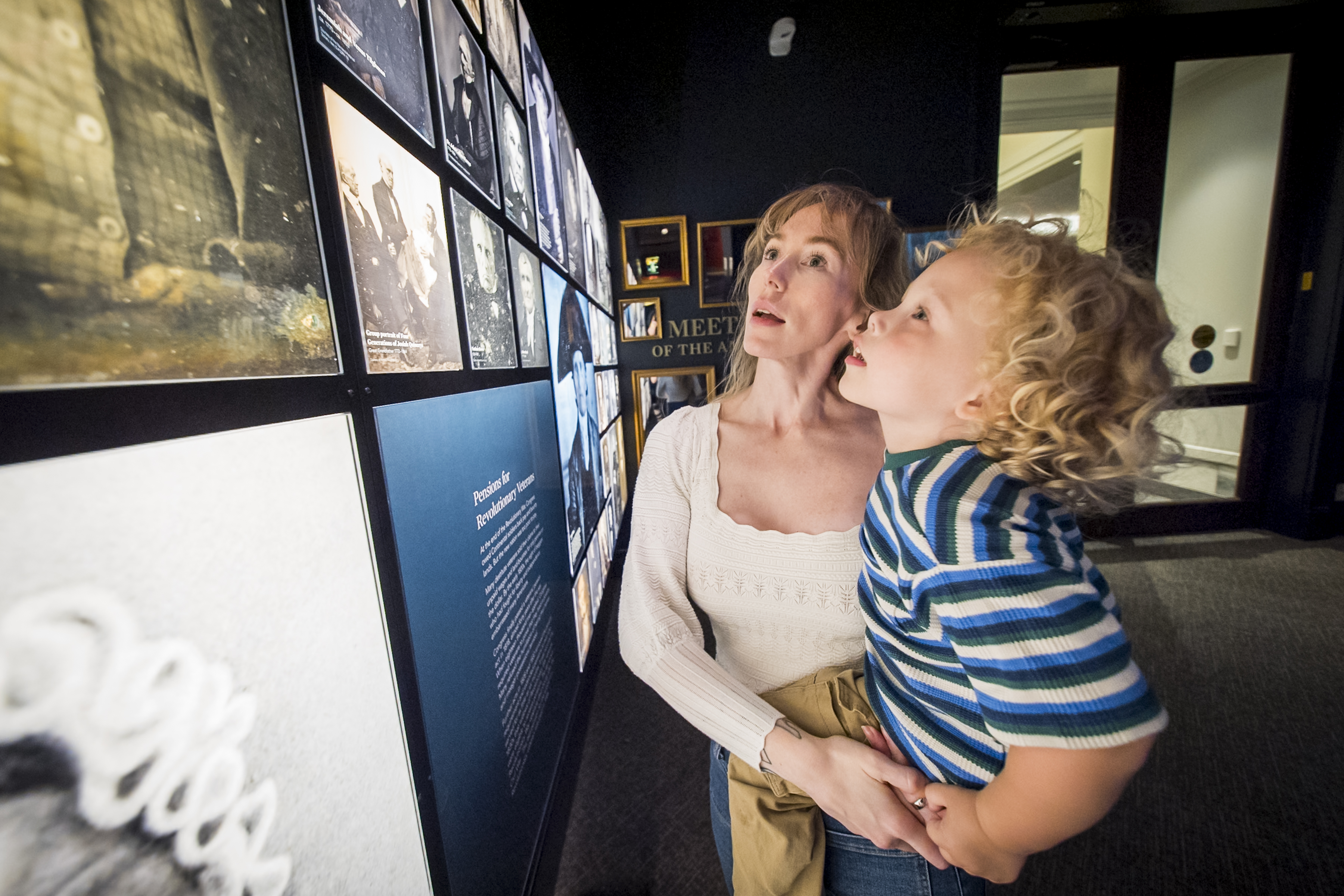 Woman holds young boy who is touching a photo wall featuring 19th century photos