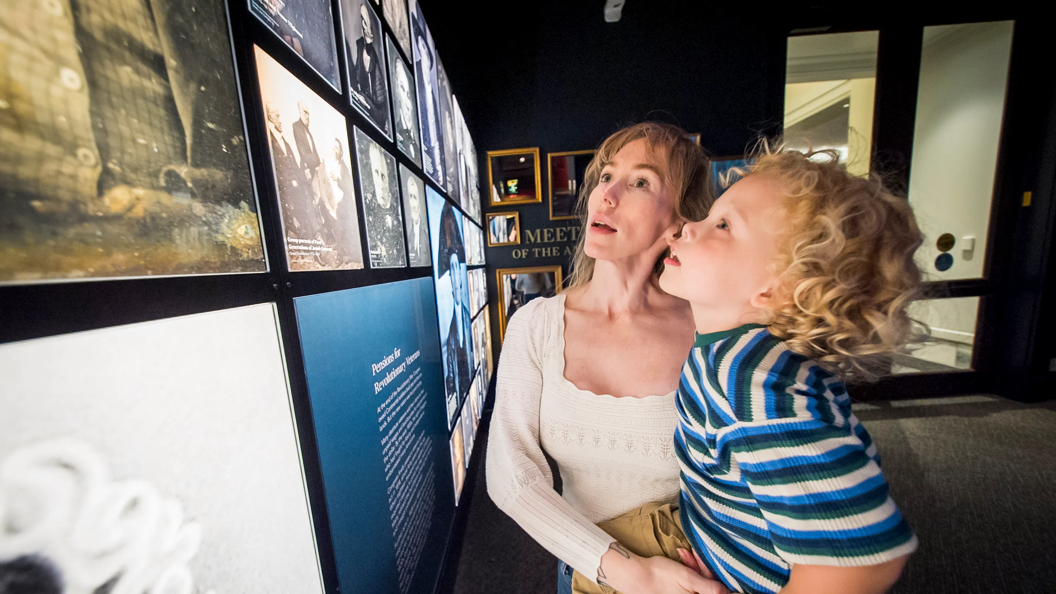 Woman holds young boy who is touching a photo wall featuring 19th century photos