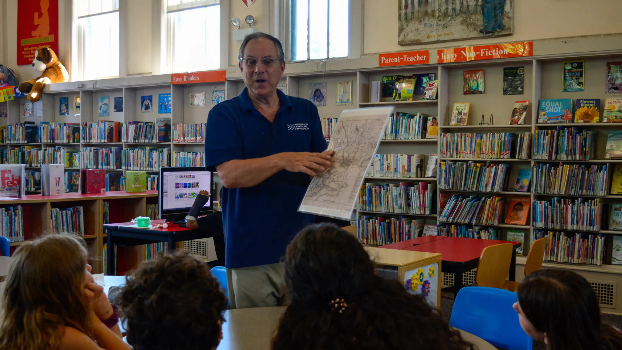 Older man with glasses in navy polo shirts points to 18th-century style map for children to see