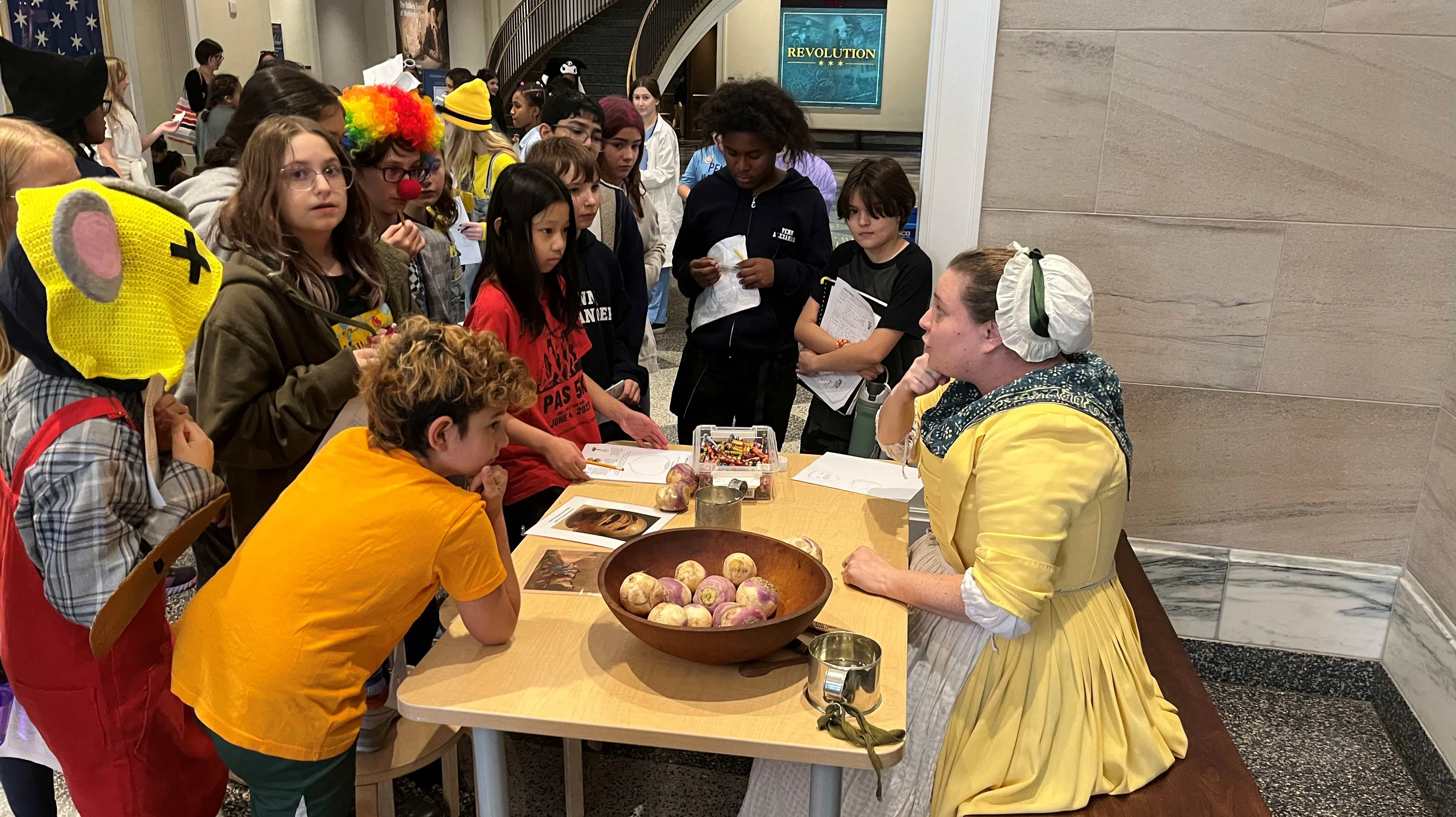 A group of guests in Halloween costumes stand in front of a table, where a costumed interpreter is sitting