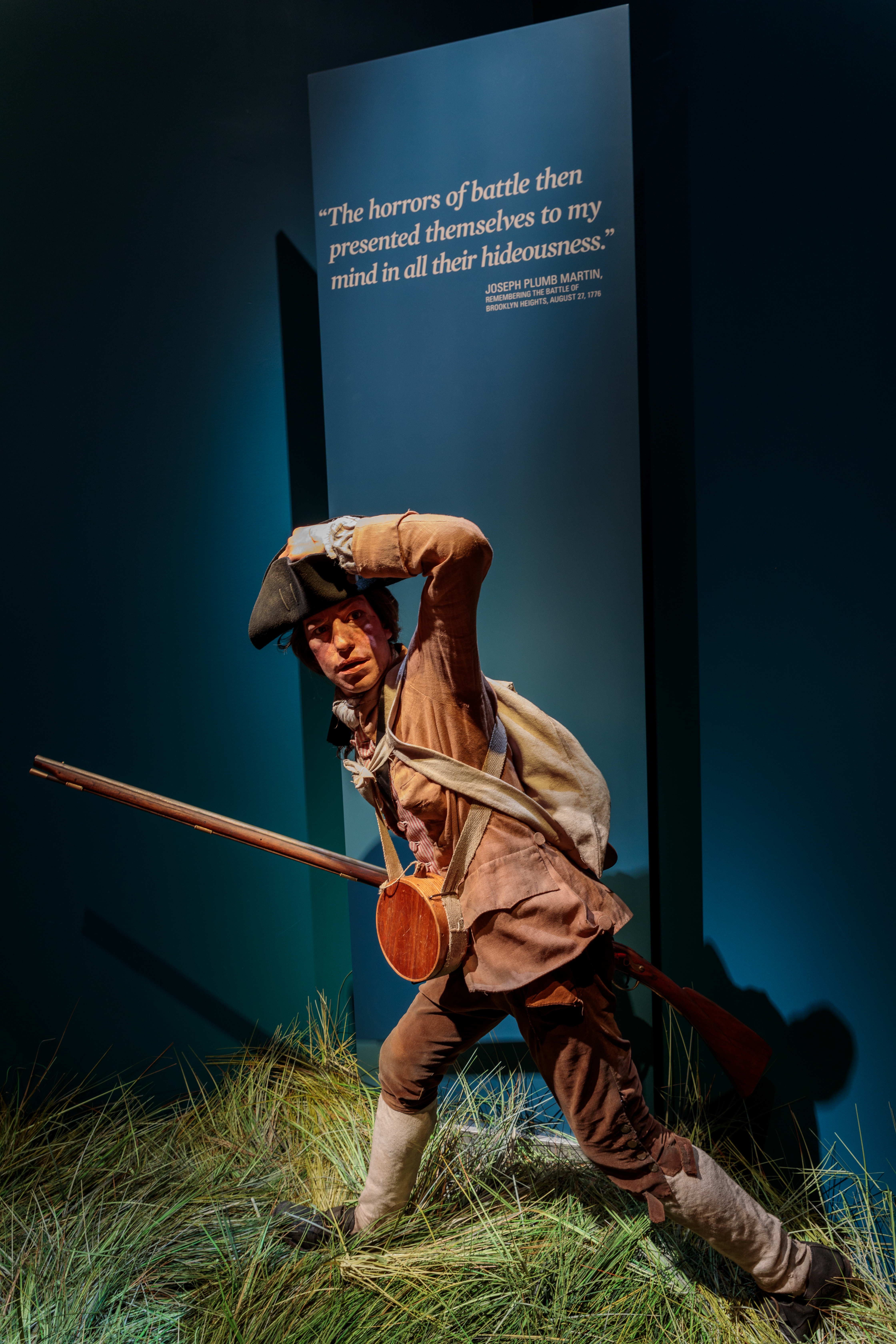 A life sized figure of a man in 18th century clothes standing on grass and carrying a rifle