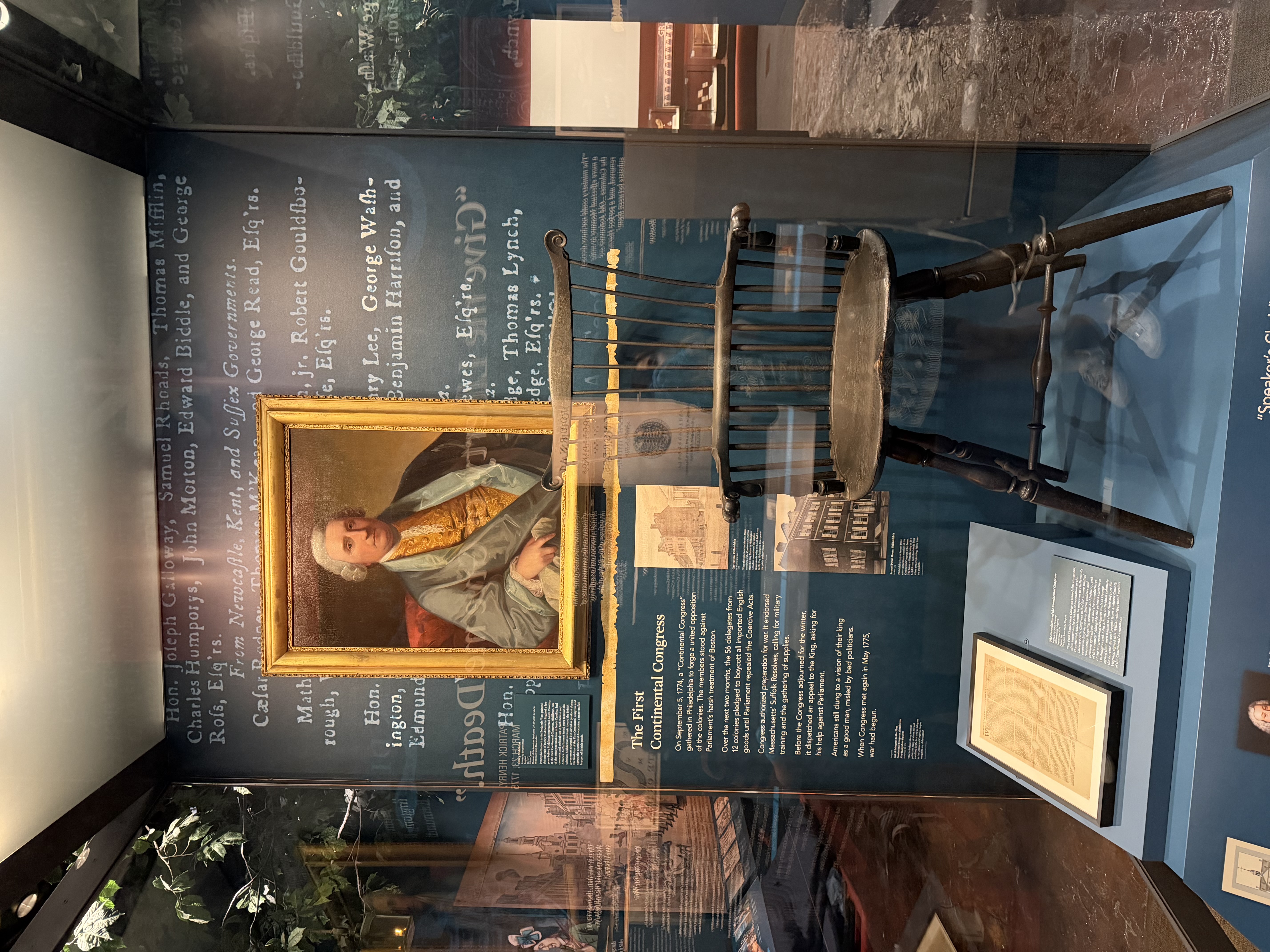 A glass case with a portrait of a white man, documents, and a wooden chair