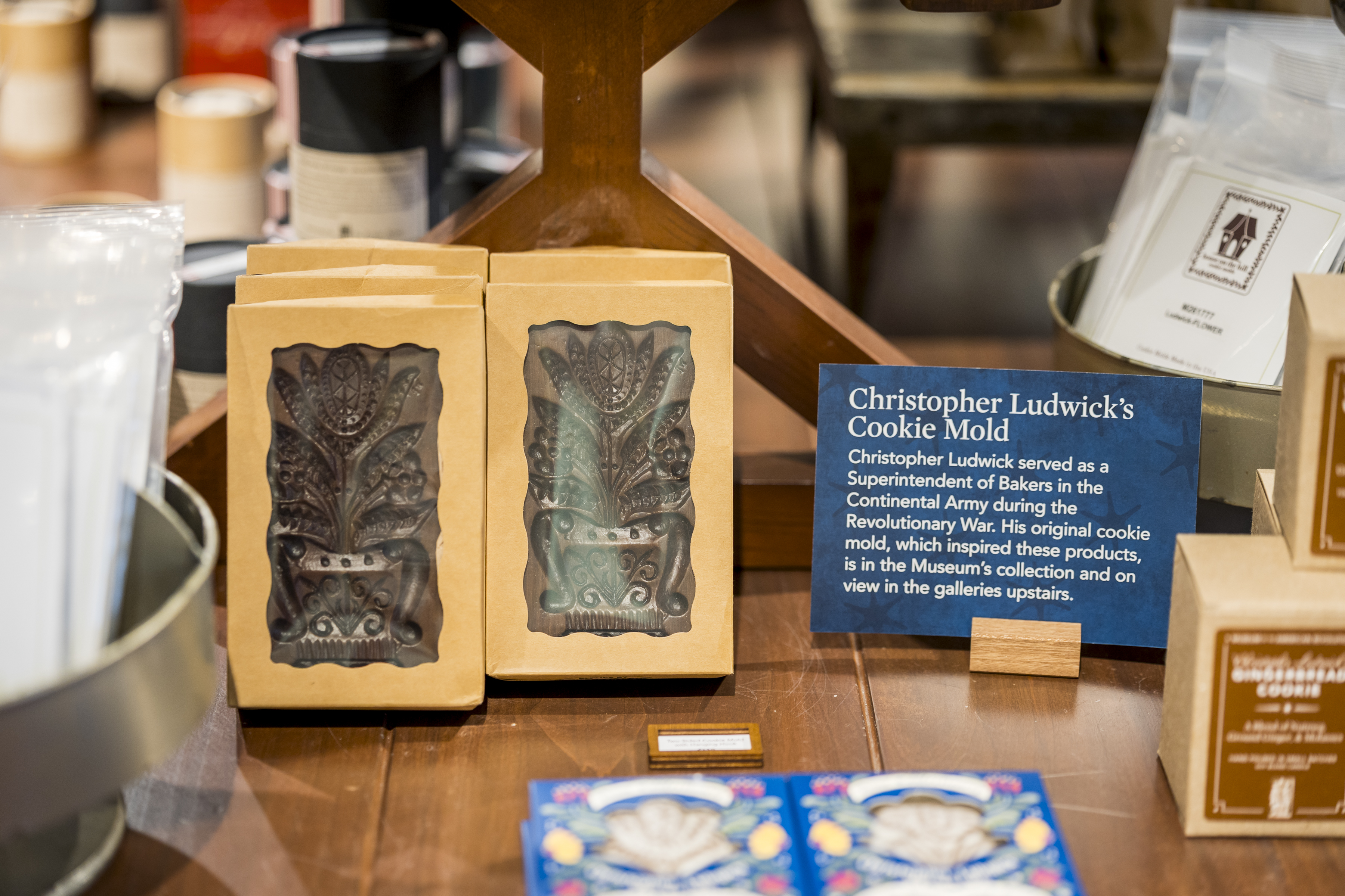 A blue sign reads "Christopher Ludwick's Cookie Mold" next to a display of brown cookie molds in tan boxes.