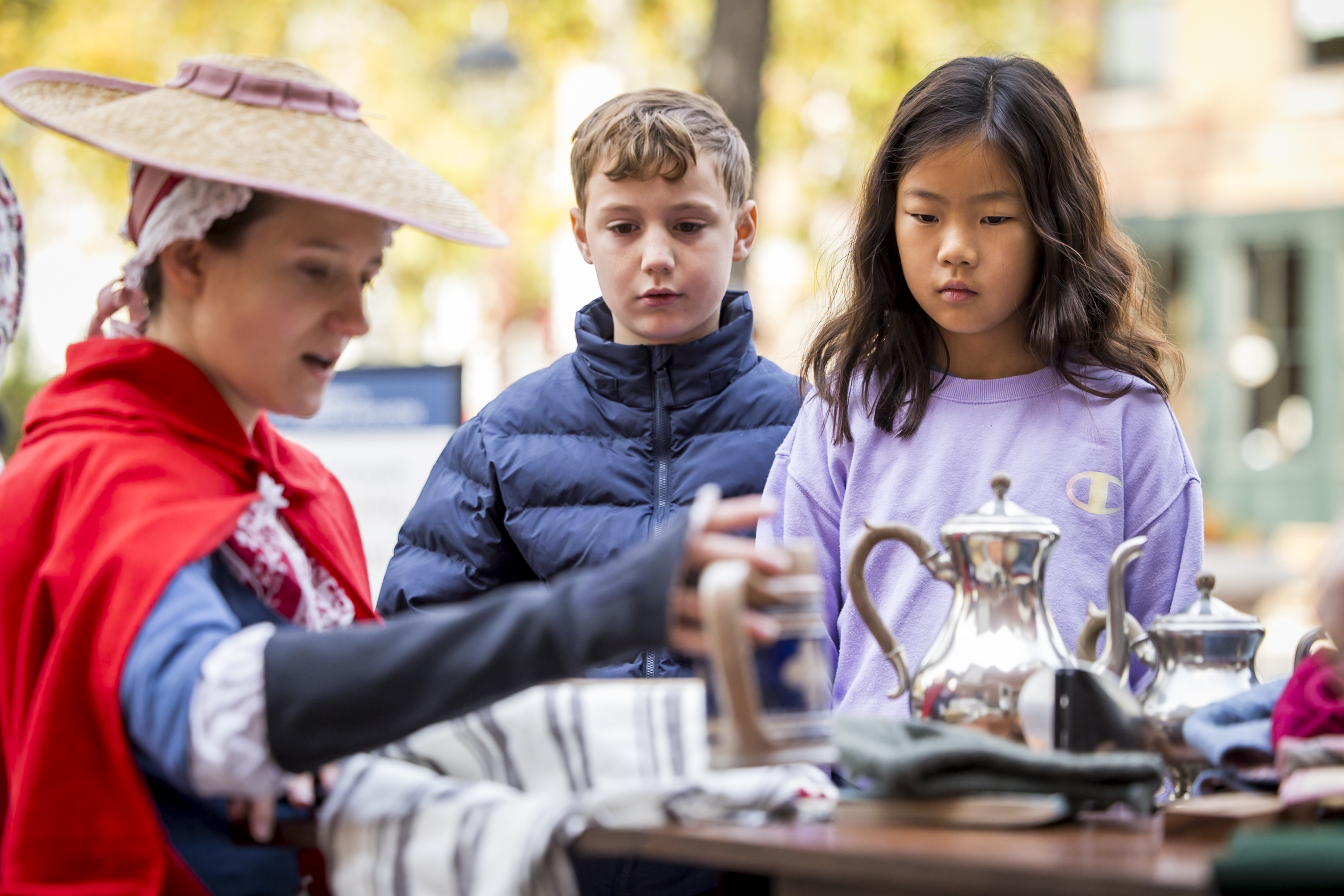 Woman in 18th-century clothing holds teapot for two kids to view