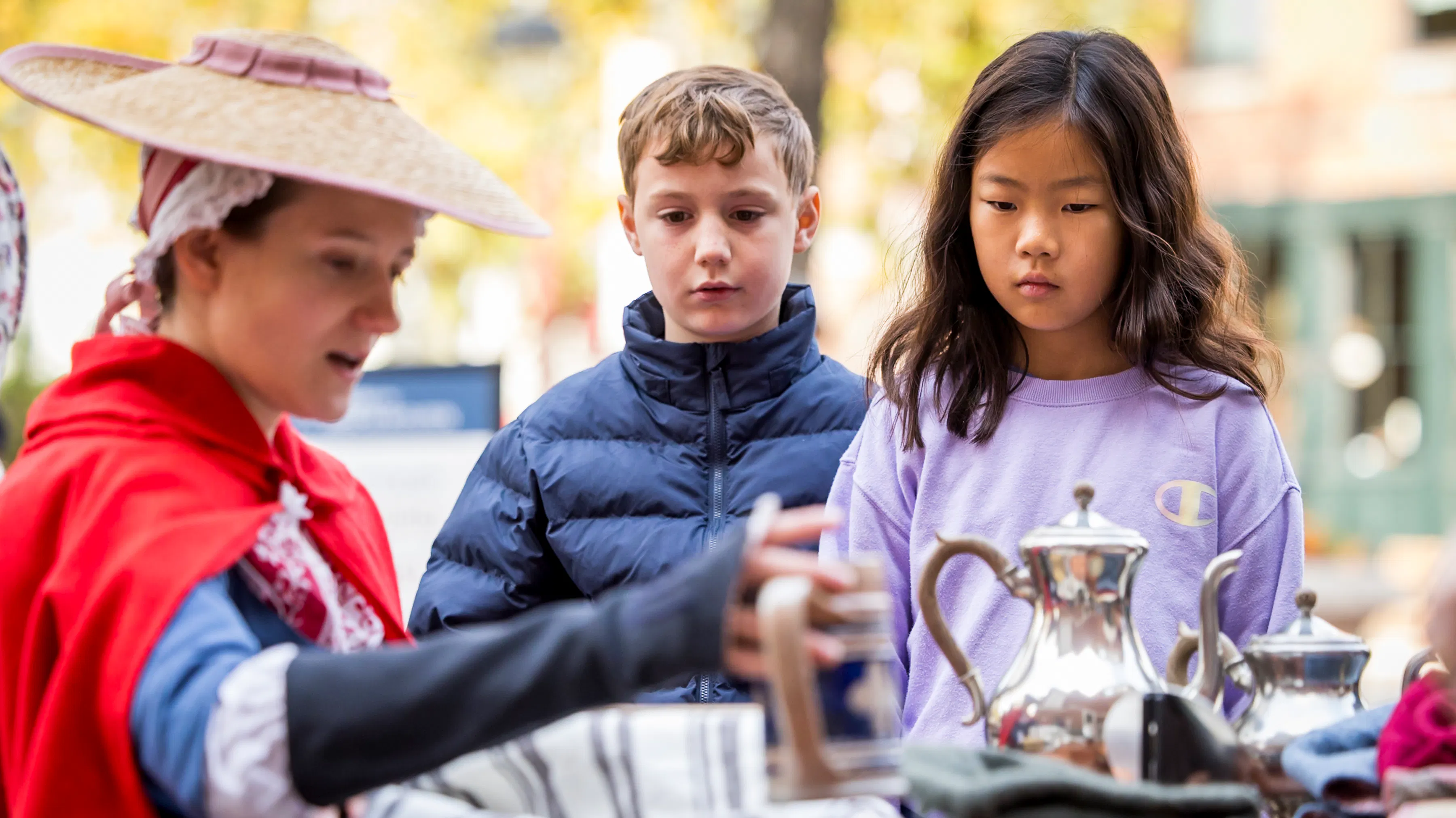 Woman in 18th-century clothing holds teapot for two kids to view