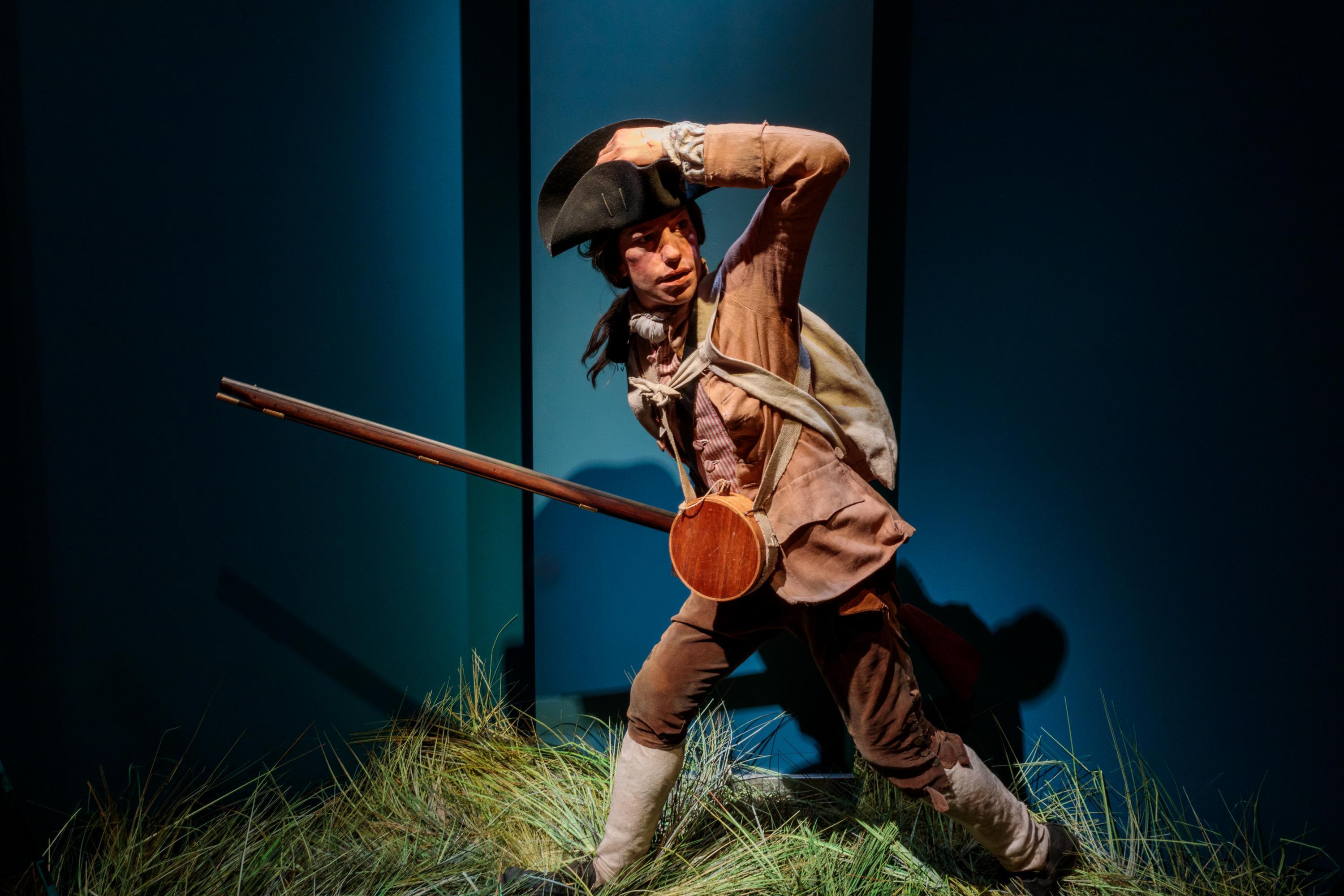 A life sized figure of a man in 18th century clothes standing on grass and carrying a rifle