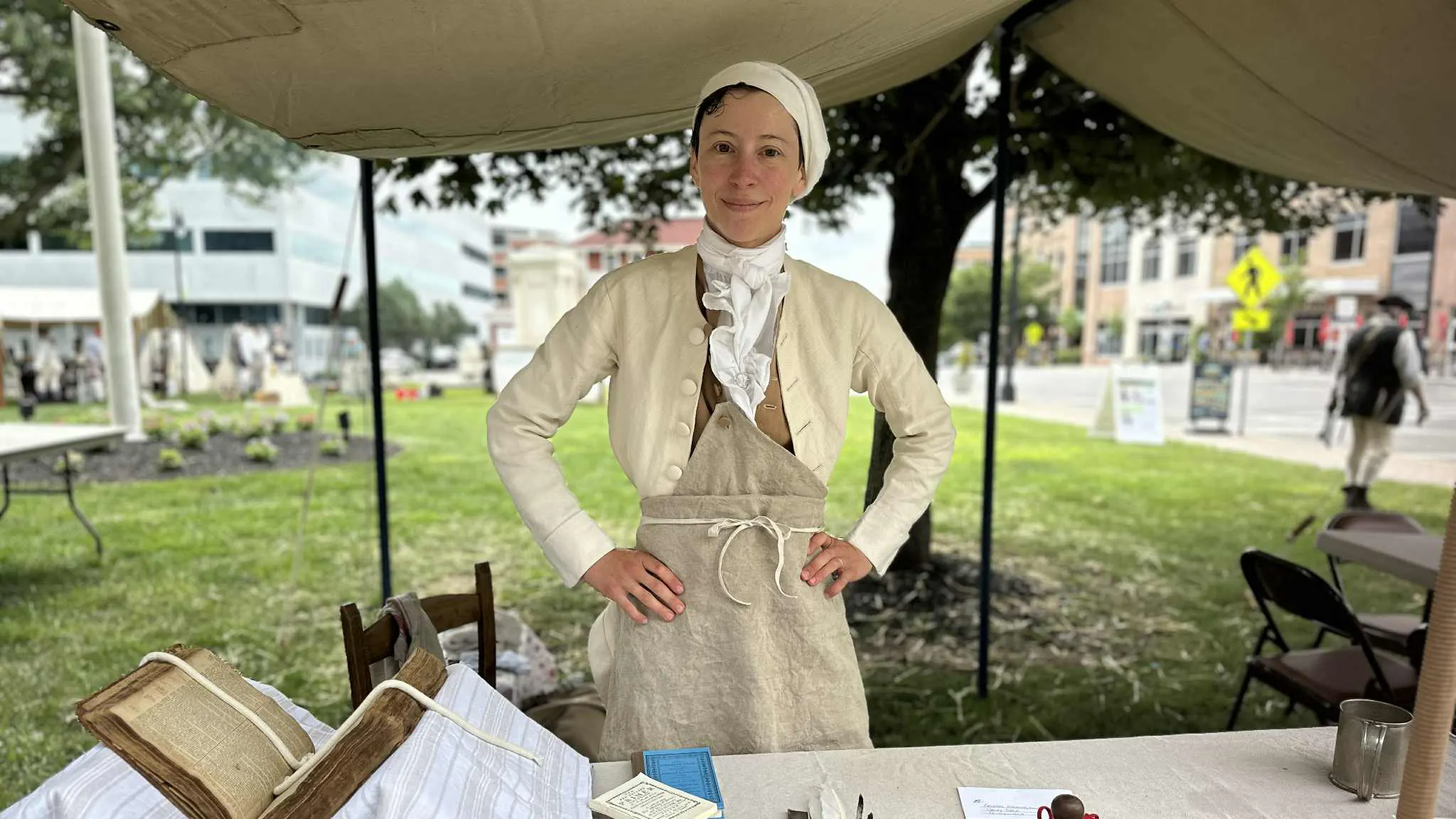 A costumed interpreter stands in an outdoor tent behind a table with bookmaking supplies