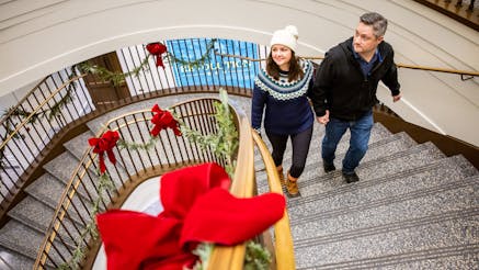 A couple holds hands while walking up the Museum's staircase decorated with green and red holiday decor.