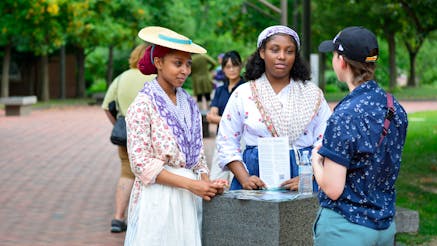 Two women in 18th century clothing speaking with person in modern clothing