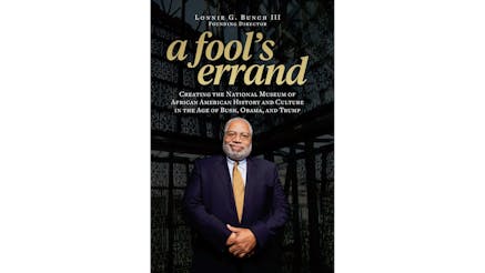This image shows the book cover of A Fools Errand: Creating the National Museum of African American History and Culture in the Age of Bush, Obama, and Trump by Lonnie Bunch, III. A Fool’s Errand is written in golden bold letters, while the subtitle and Lonnie’s name are written in white font. There is a photograph of Lonnie in a suit and his hands folded in front of him. He is smiling at the viewer.