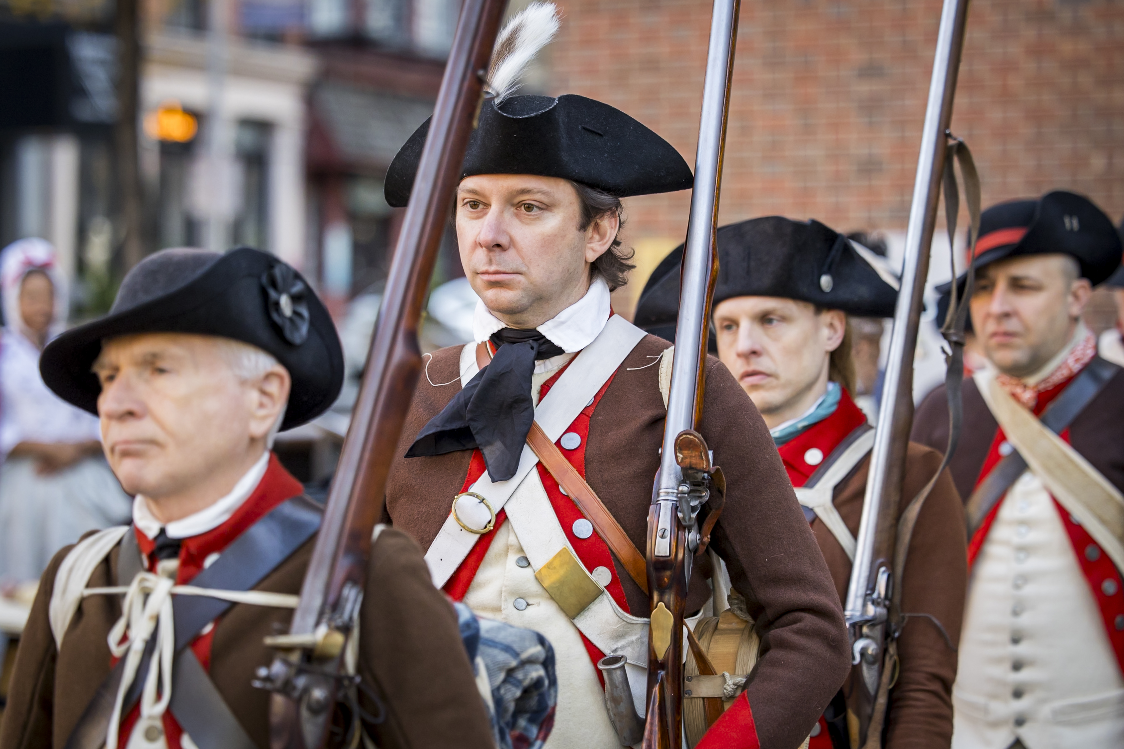 Four 18th-century American soldiers in brown coats and black hats marching with guns on their shoulders