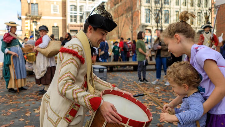 A costumed interpreter lets two young children play a drum on the Museum's plaza.