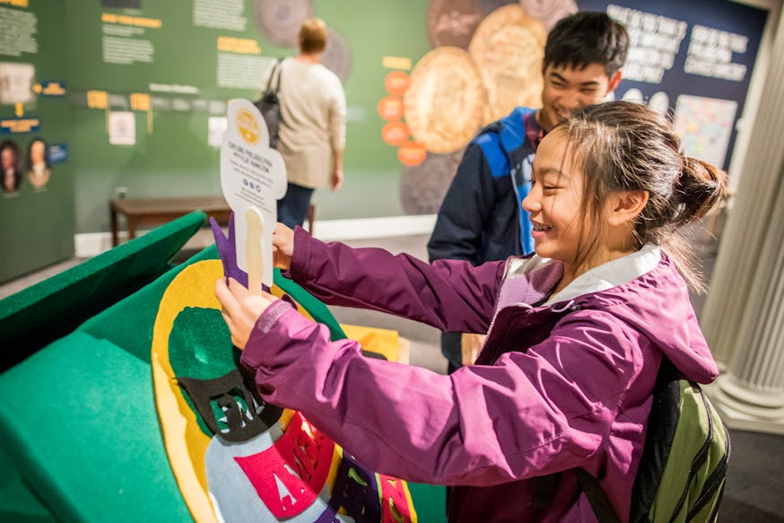 Two Asian American youths, one male and one female, interact with a child friendly activity in the Hamilton Was Here exhibit. The female holds the Flat Hamilton in her left hand, with the back of it facing the viewer. They are both smiling. In the background, a woman, with her back to the viewer, reads the exhibit panels.