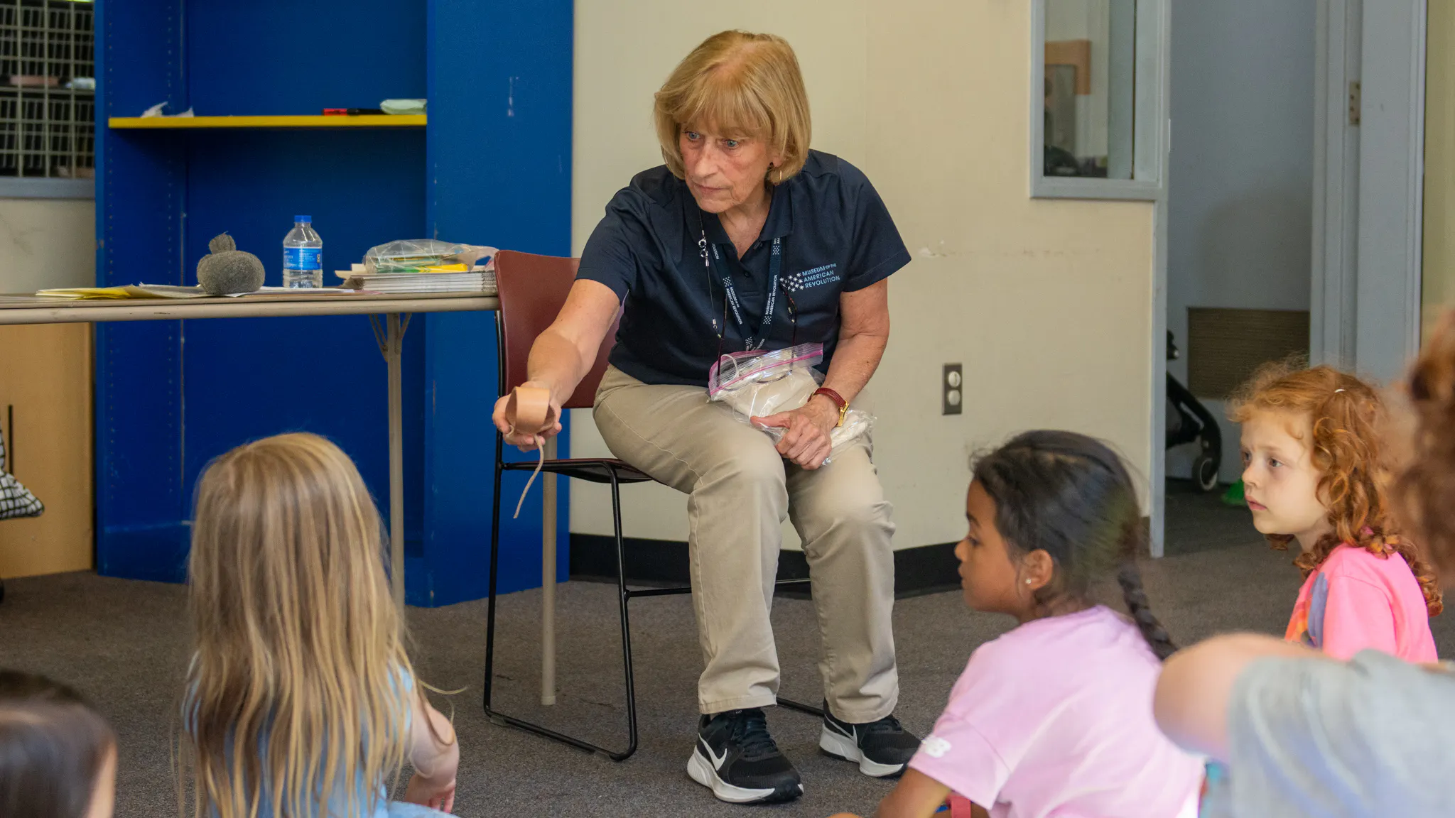Woman with light blonde hair, navy polo, and khakis speaks to group of children 03credit  Moar