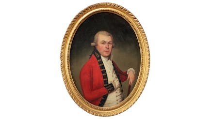This image shows a gold framed portrait of Thomas McDonogh. The portrait is against a white background. Thomas is wearing a red jacket with a blue shirt. His right hand is resting inside a buttonhole of the shirt, so you cannot see his hand. His left wrist is placed on the wooden chair he is sitting on, with his hand dangling over. His eyebrows are raised as he stares at the viewer.