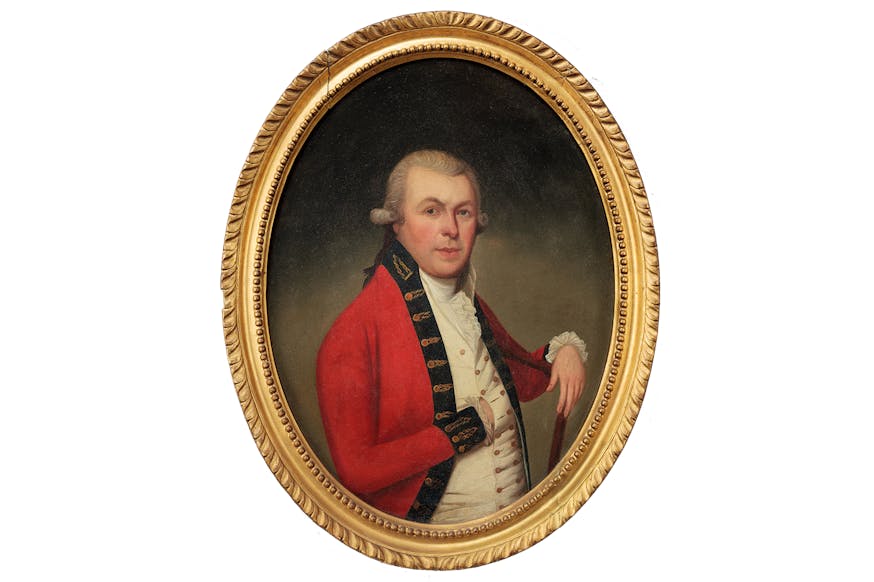 This image shows a gold framed portrait of Thomas McDonogh. The portrait is against a white background. Thomas is wearing a red jacket with a blue shirt. His right hand is resting inside a buttonhole of the shirt, so you cannot see his hand. His left wrist is placed on the wooden chair he is sitting on, with his hand dangling over. His eyebrows are raised as he stares at the viewer.
