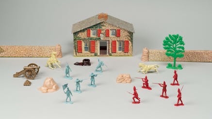 Blue and red toy soldier pieces in front of a toy recreated 18th century house.