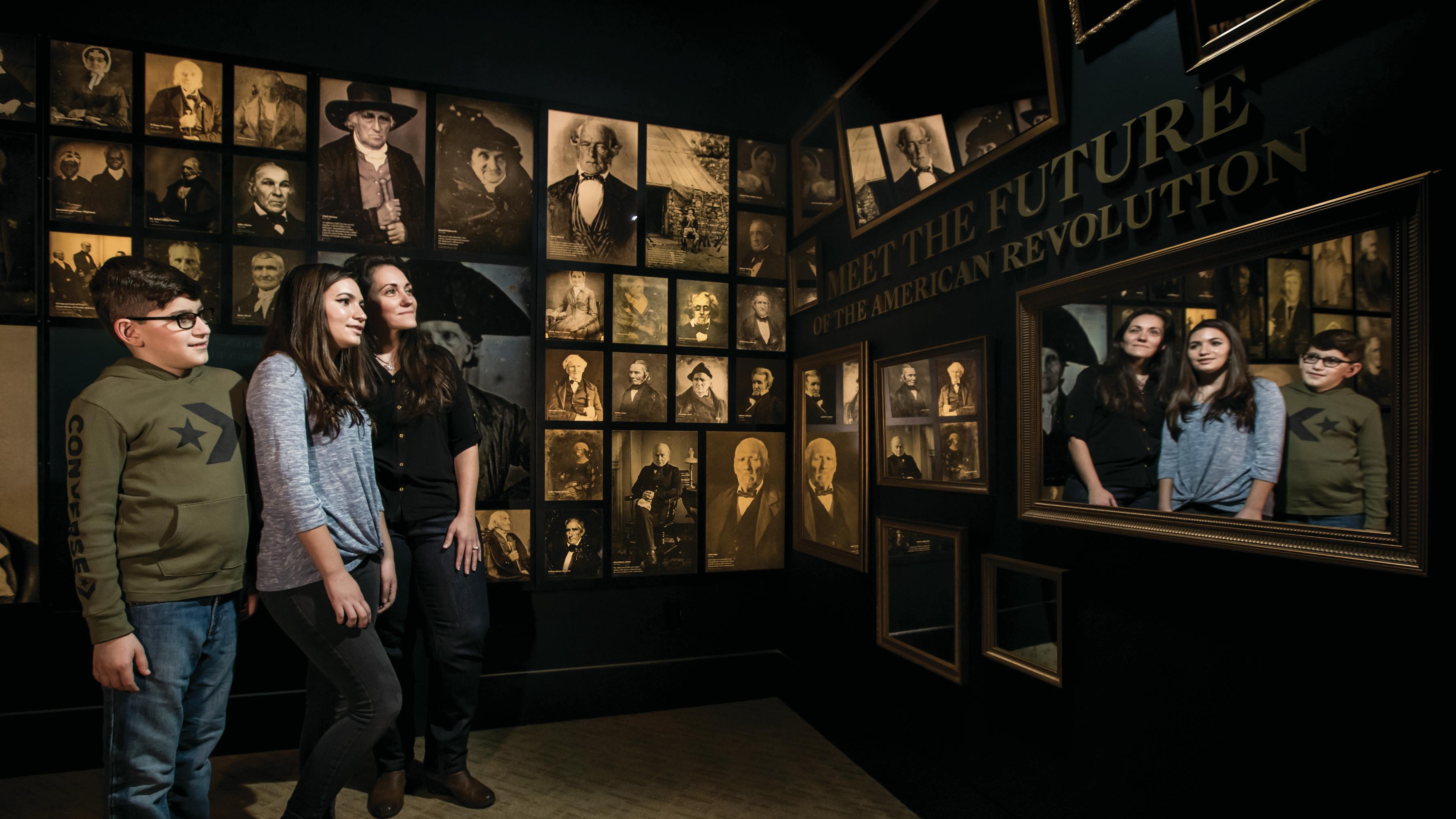 Three guests stand in front of a mirror on the Museum's Meet the Revolutionary Generation wall. This is a gallery of photographs that show people who lived through the Revolutionary War.