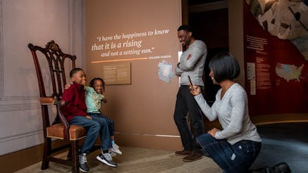 A mother takes a photo of her son and daughter sitting on a chair in the Writing the Constitution section inside the Museum galleries. She is bended on one knee as her husband stands next to her.