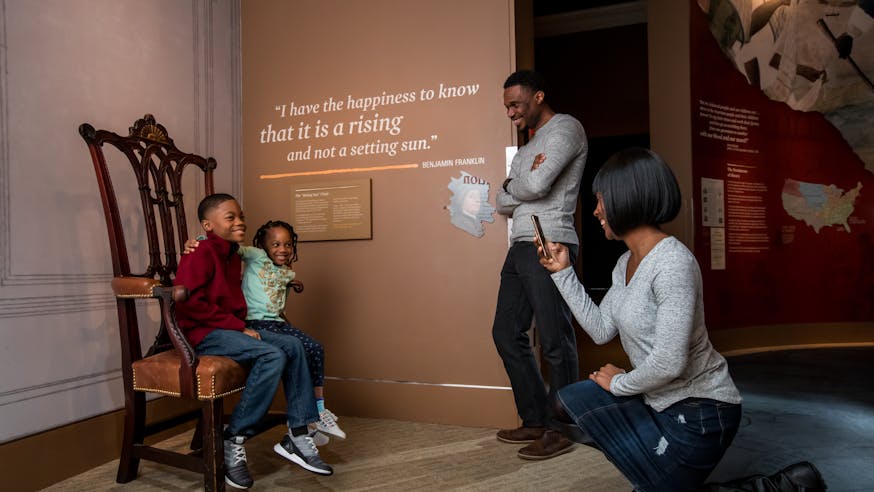 A mother takes a photo of her son and daughter sitting on a chair in the Writing the Constitution section inside the Museum galleries. She is bended on one knee as her husband stands next to her.