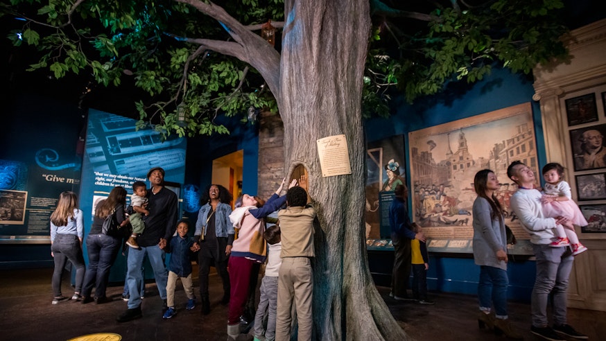 The Boston Liberty Tree is in the middle of the Becoming Revolutionaries gallery. Guests explore the gallery.