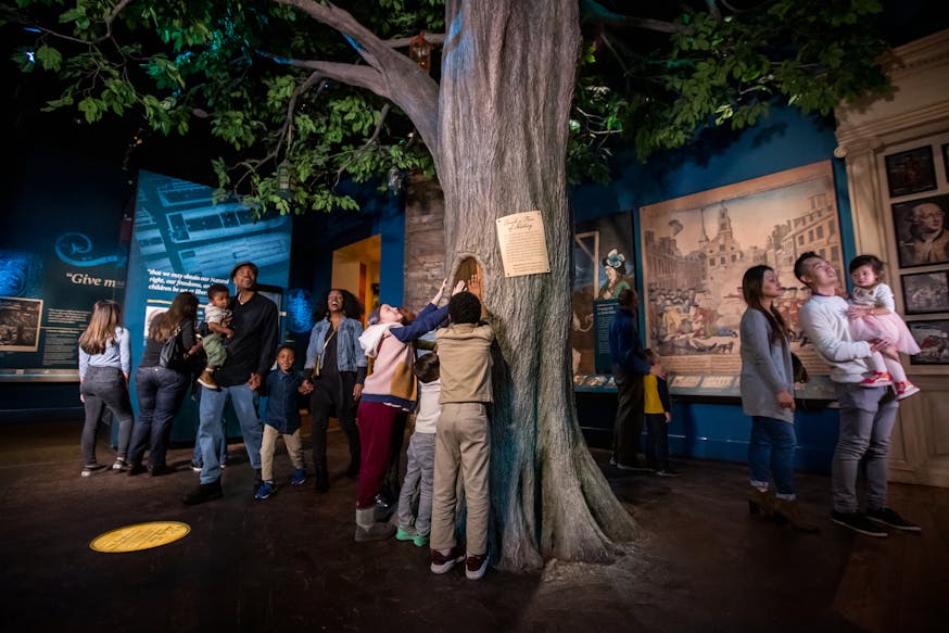 The Boston Liberty Tree is in the middle of the Becoming Revolutionaries gallery. Guests explore the gallery.