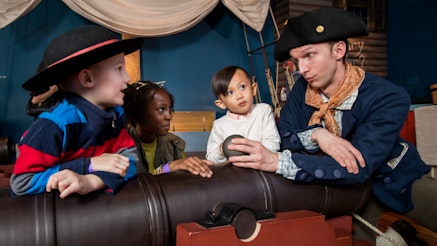 A Museum educator, in Revolutionary military costume, shows three adolescents how to use a cannon on the Privateer Ship in the galleries.