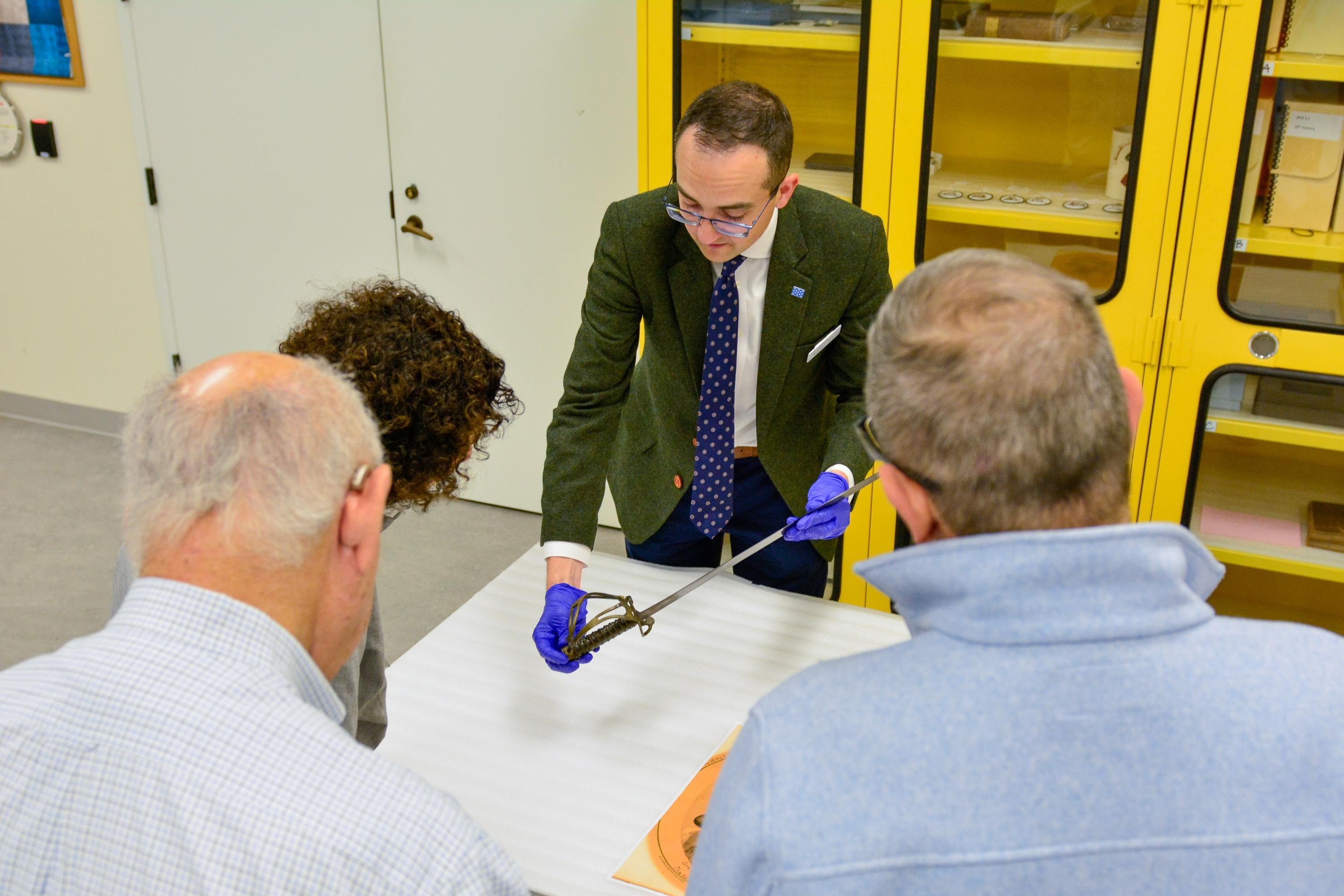 A man standing behind a table holds a sword for three people to view
