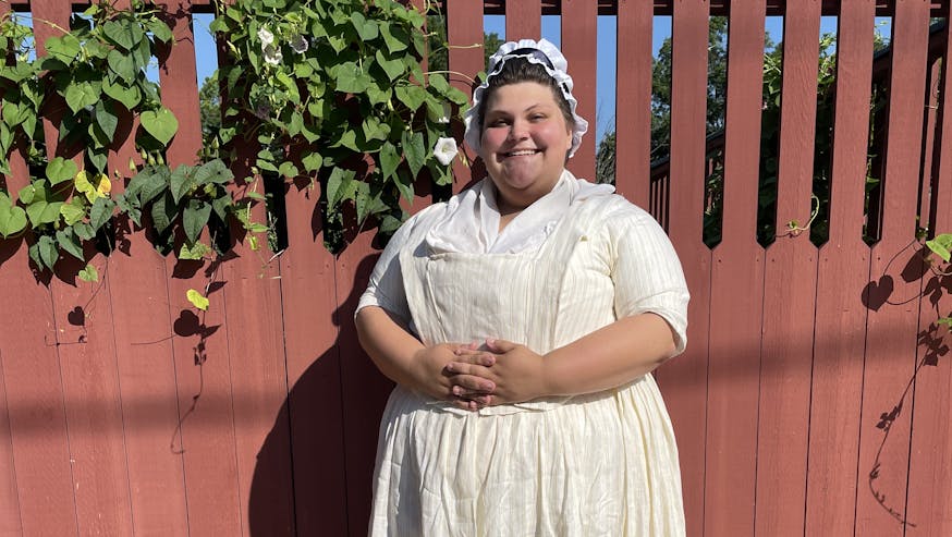 Historical interpreter Sydney Marenburg dressed in a white 18th century dress and standing in front of a red fence with green vines.