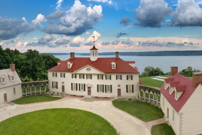 The Mount Vernon estate with green lawn in front and beige mansion with red roof and Potomac River in the background.