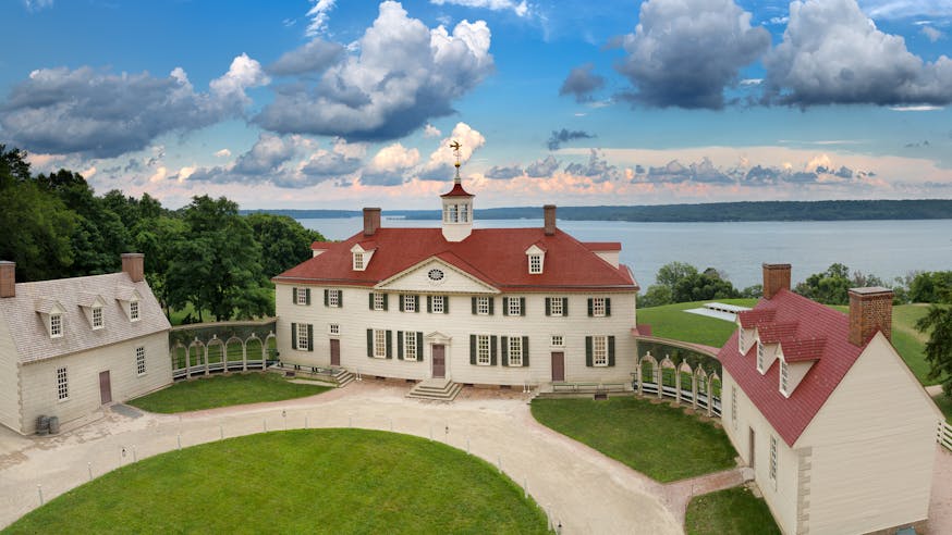The Mount Vernon estate with green lawn in front and beige mansion with red roof and Potomac River in the background.