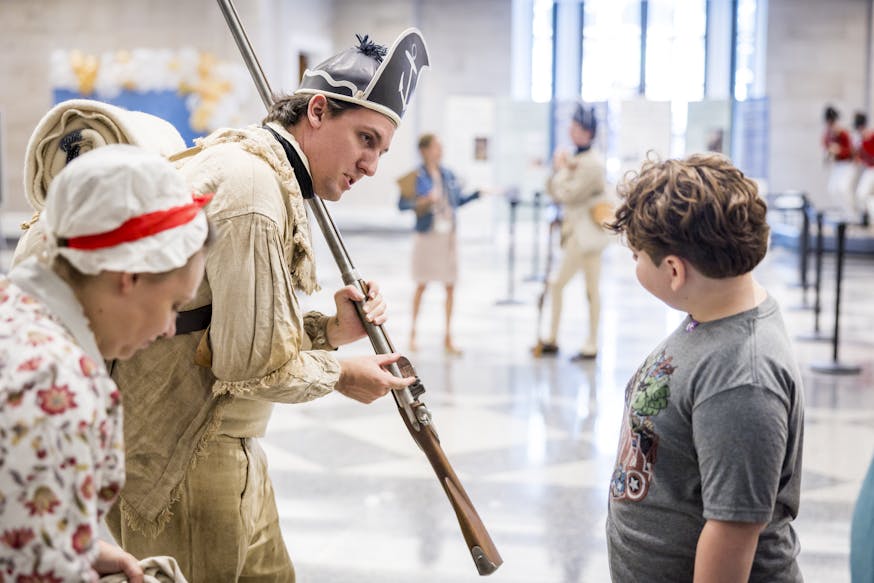 Costumed living historian Hayden Conley wears a beige hunting shirt and black cap while showing part of a musket to a young visitor inside the Museum's rotunda.