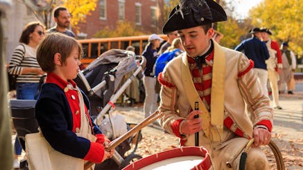 A British costumed living history interpreter shows a young boy how to drum on the Museum's plaza during Occupied Philadelphia.