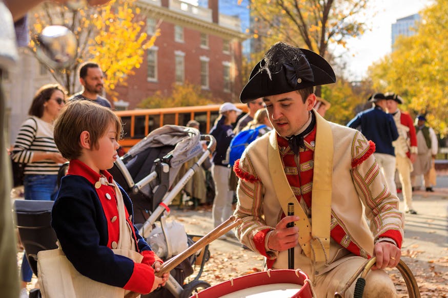 A British costumed living history interpreter shows a young boy how to drum on the Museum's plaza during Occupied Philadelphia.