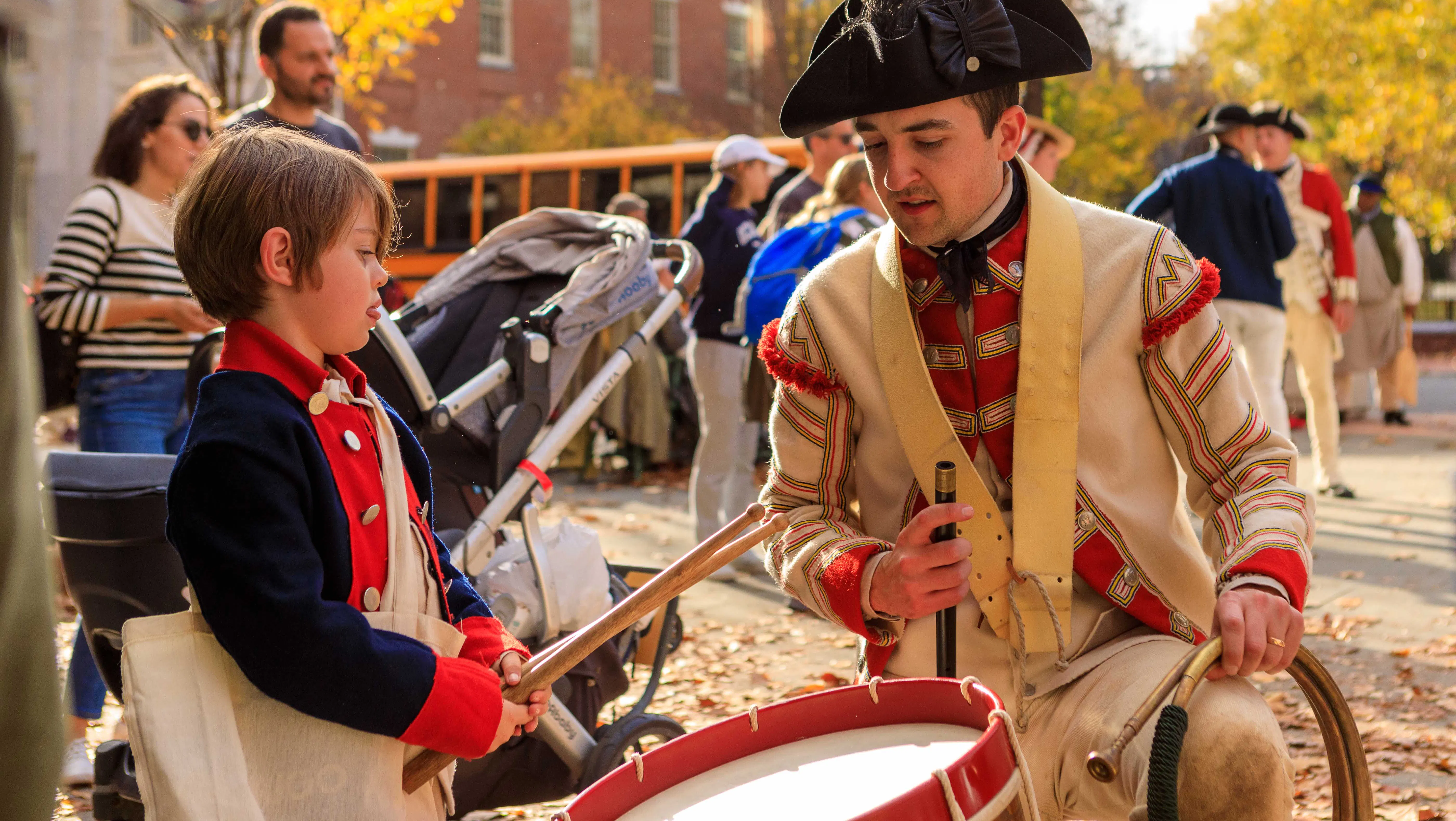 A British costumed living history interpreter shows a young boy how to drum on the Museum's plaza during Occupied Philadelphia.