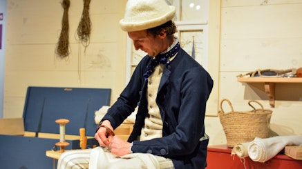 A costumed living history interpreter demonstrates sewing a sail.